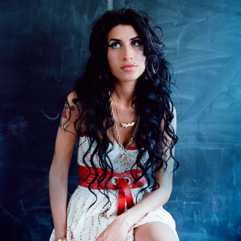 Amy Winehouse wearing a white and red dress against a blue backdrop in a promotional image for the Back to Black 20th anniversary editions.