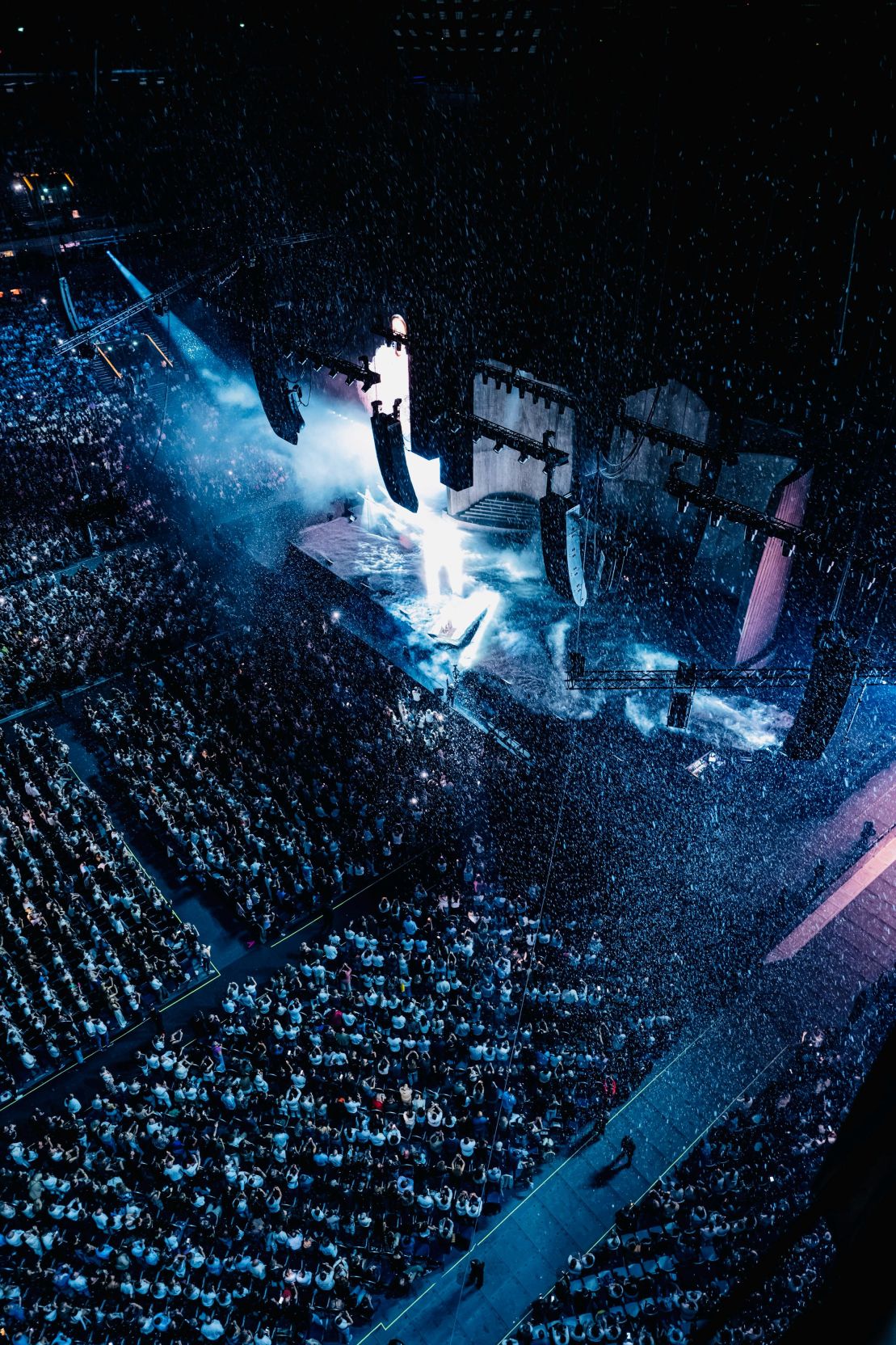 Overhead view of a packed Plenitude Arena with blue stage lighting and falling confetti during Céline Dion’s 2026 Paris concerts.