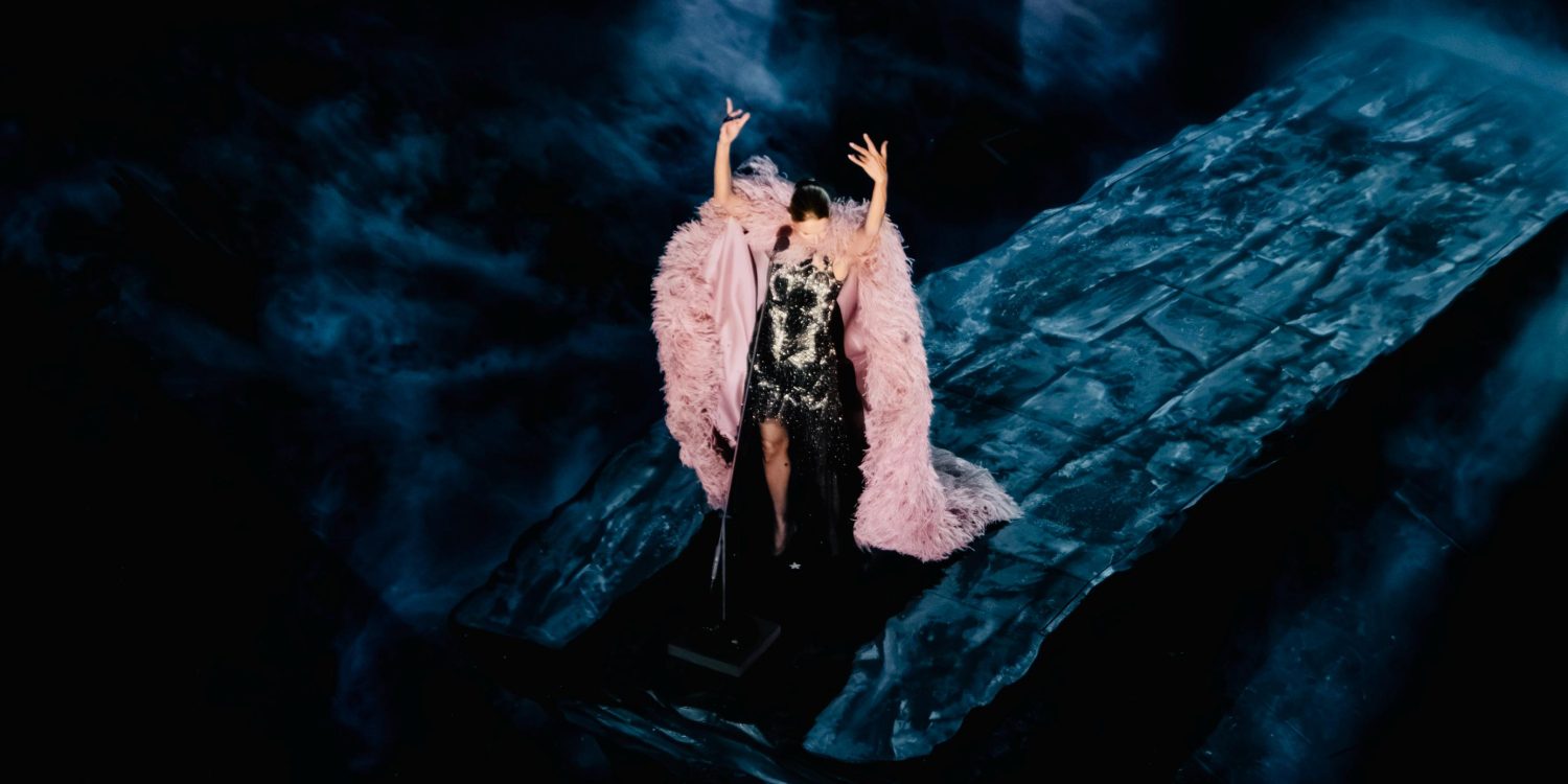 Céline Dion raises her arms in a pink feathered cape on a blue-lit stage during her 2026 Paris concerts.
