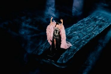 Céline Dion raises her arms in a pink feathered cape on a blue-lit stage during her 2026 Paris concerts.