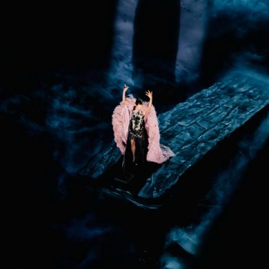 Céline Dion raises her arms in a pink feathered cape on a blue-lit stage during her 2026 Paris concerts.