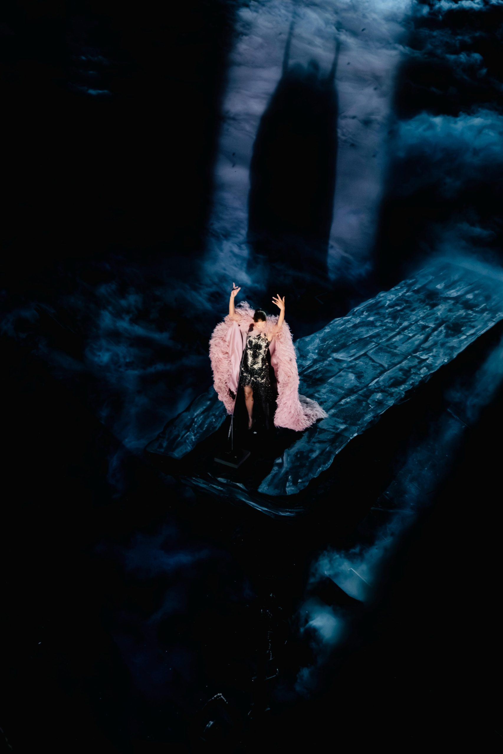 Céline Dion raises her arms in a pink feathered cape on a blue-lit stage during her 2026 Paris concerts.