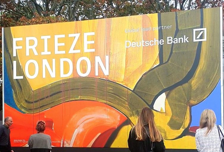 Visitors stand before the colourful Frieze London entrance sign beneath autumn trees in Regent’s Park.
