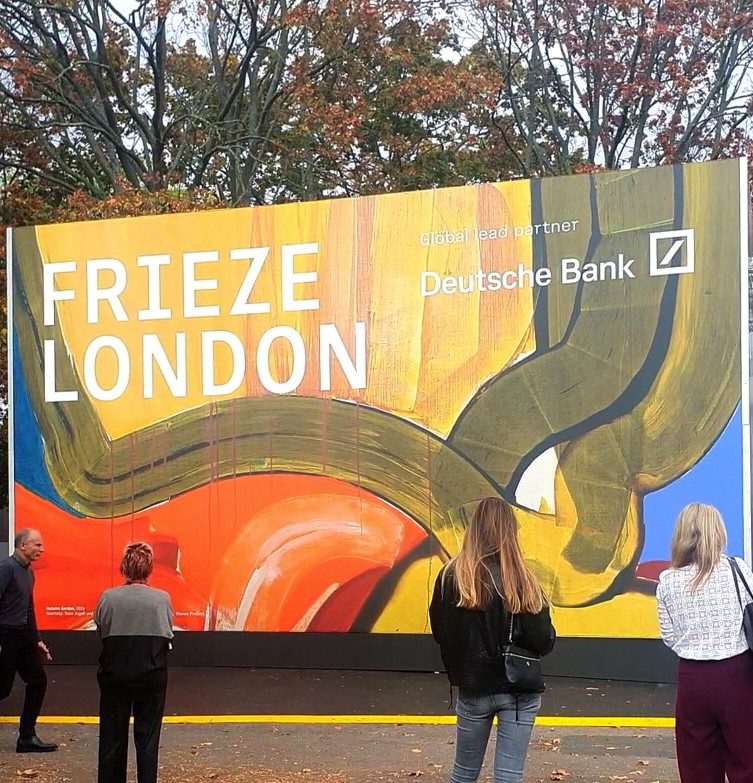 Visitors stand before the colourful Frieze London entrance sign beneath autumn trees in Regent’s Park.