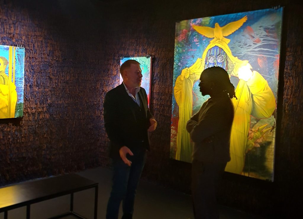 Ken Nwadiogbu in conversation with a visitor beside his yellow masquerade paintings in the Degussa Lounge.