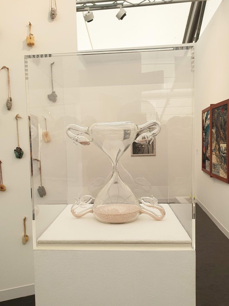 Lea Cetera’s glass hourglass with uterus-like forms and pale sand, displayed inside a clear case on a white plinth.