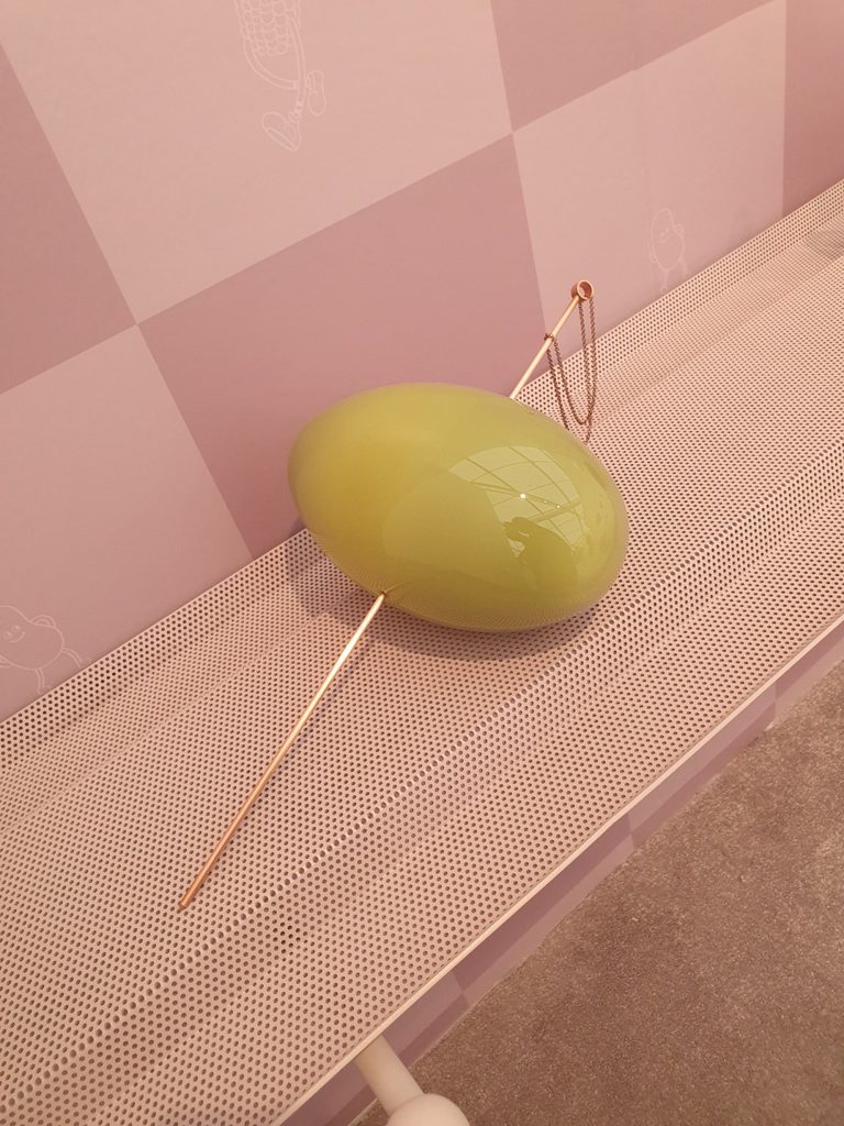 Oversized green glass olive pierced by a metal skewer on a perforated shelf against a pink checked wall.