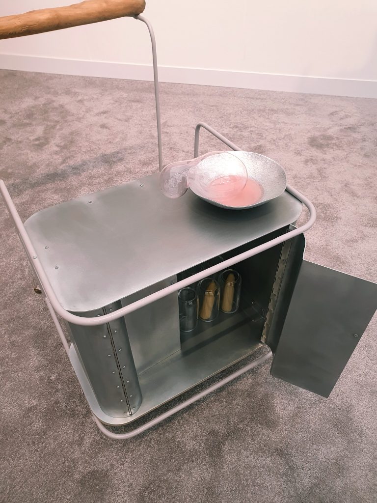 Pink liquid and a tipped glass vessel in a metal bowl on a serving trolley in Steph Huang’s installation.