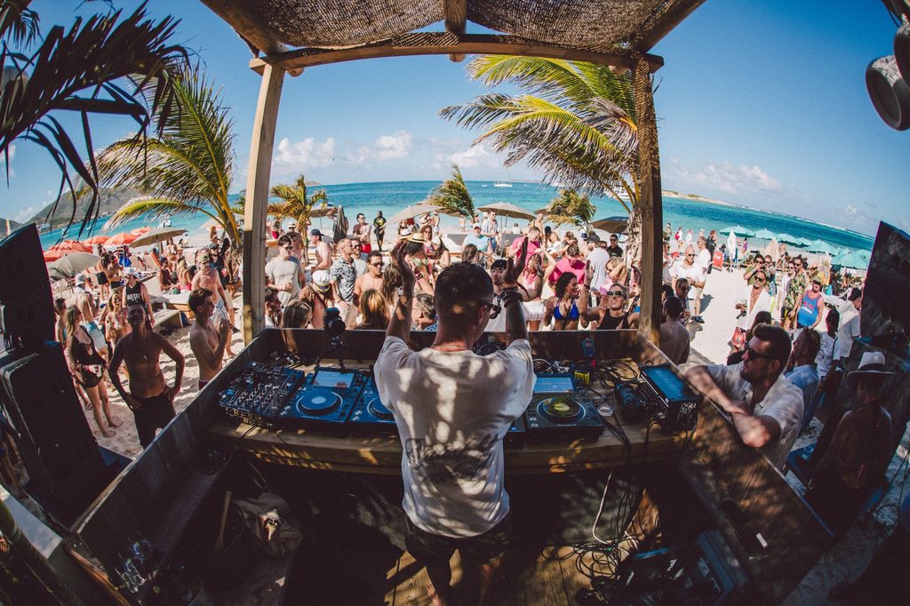 DJ performing to a crowd at an SXM Festival beach party in St. Martin, with palm trees and the Caribbean Sea in the background
