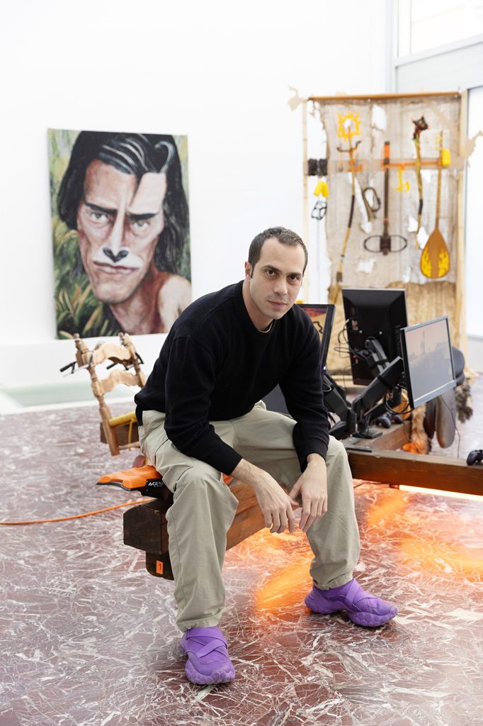 Theo Triantafyllidis seated in his art studio wearing purple trainers, with colourful artworks and creative equipment behind him.